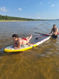 На фотографии изображены два человека на сапборде в воде. Женщина в красном купальнике лежит на животе на передней части доски, а мужчина в красных плавках сидит на задней части доски и держит весло. Вода вокруг них спокойная и прозрачная. На заднем плане виден берег с лесом, а также несколько человек, находящихся в воде. Вдалеке видны две большие трубы, из которых идет дым, и другие промышленные сооружения. Небо ясное с небольшим количеством облаков.